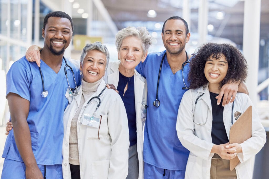 Five clinicians in scrubs and white coats standing together, smiling Five clinicians in scrubs and white coats standing together, smiling